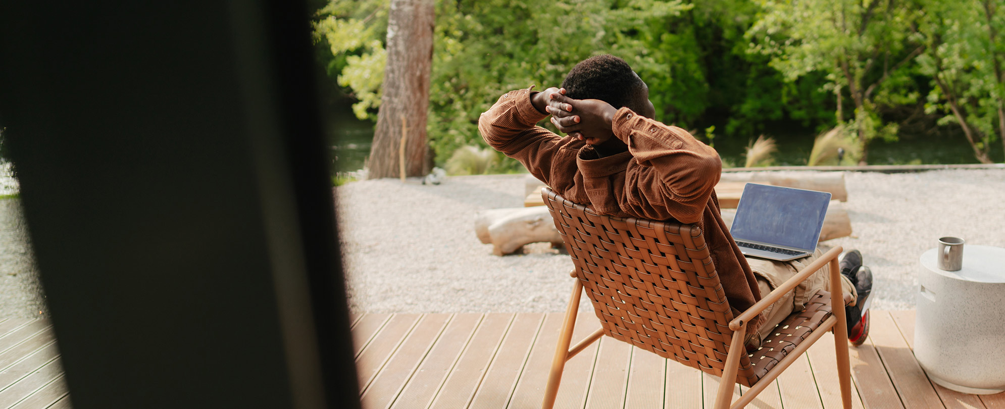 A man leans back while sitting on a patio with a laptop on his lap and a mug on a nearby table