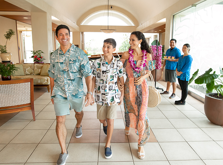 A mom, dad, and son walk through a Club Wyndham resort lobby smiling and wearing leis on their summer vacation.